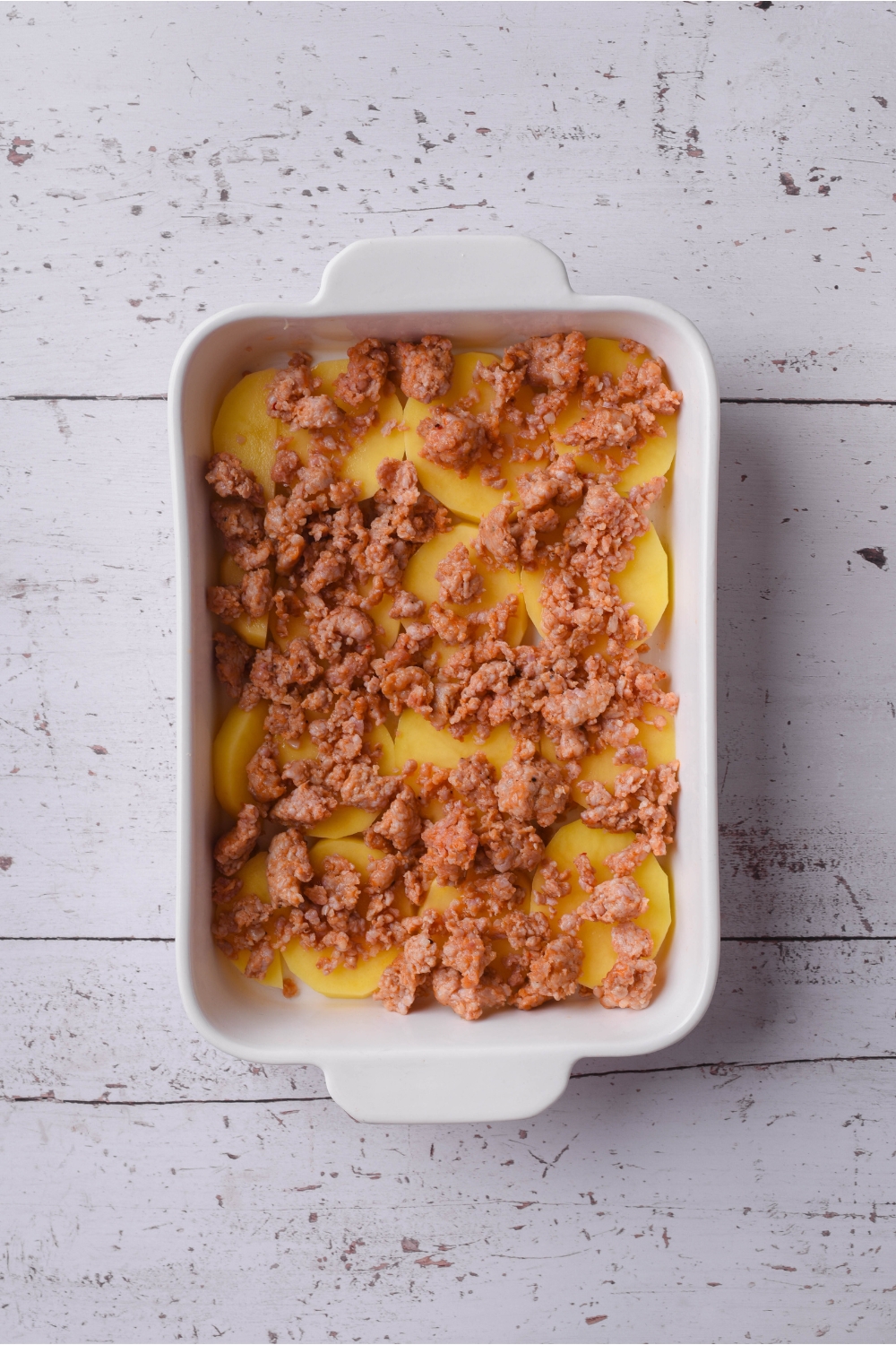A baking dish filled with a layer of potato slices and cooked sausage on top of the potatoes.