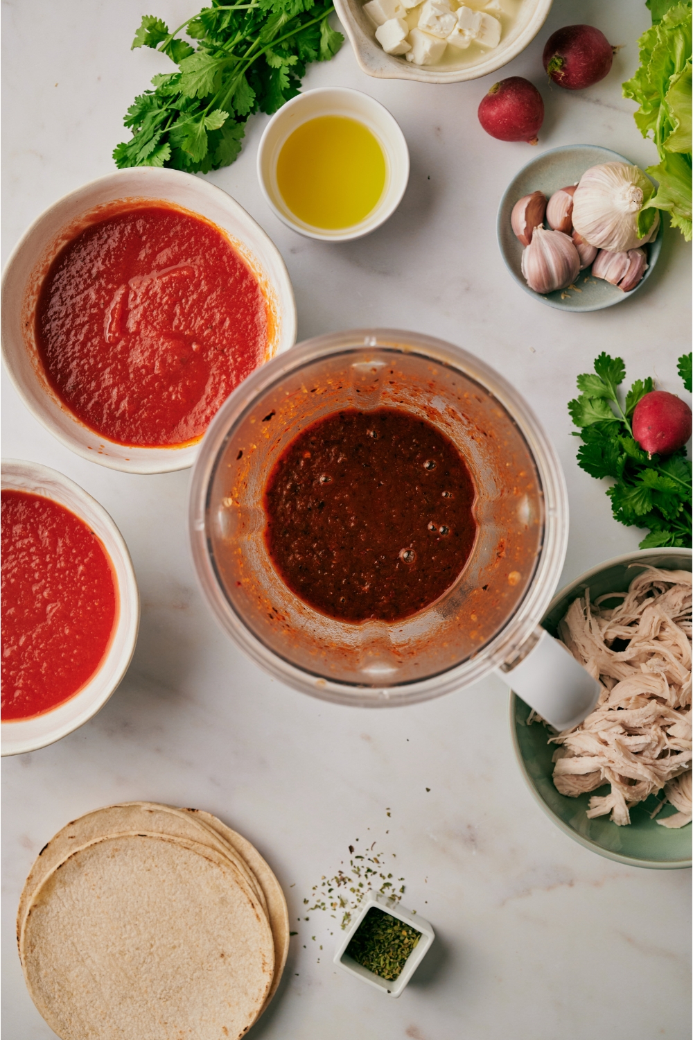 A blender filled with a chili sauce mixture surrounded by an assortment of ingredients including bowls of tomato sauce, shredded chicken, garlic cloves, and a stack of tortillas.