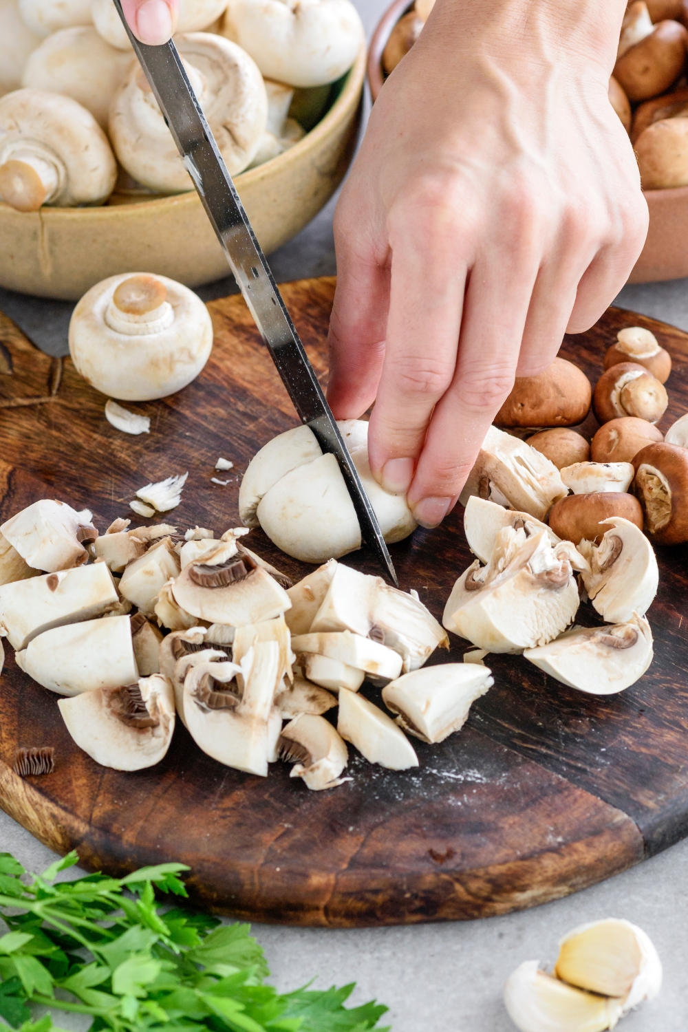 A hand chopping a mushroom into four pieces. The mushroom is on a cutting board surrounded by sliced mushrooms.