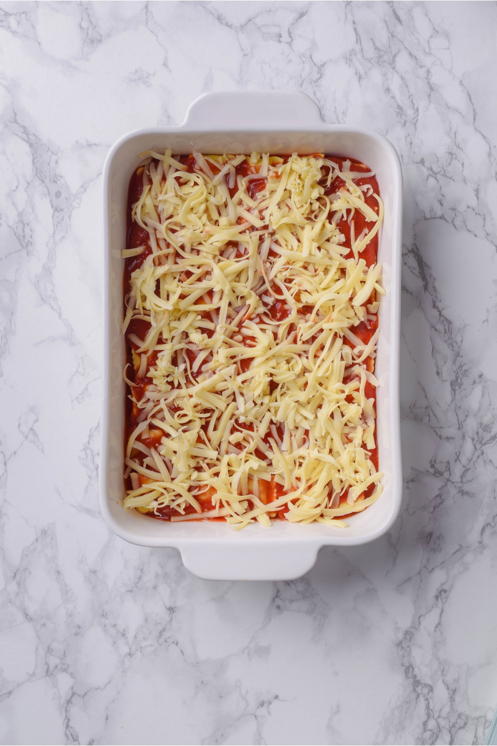 A baking dish filled with unbaked ravioli casserole topped with shredded mozzarella cheese.