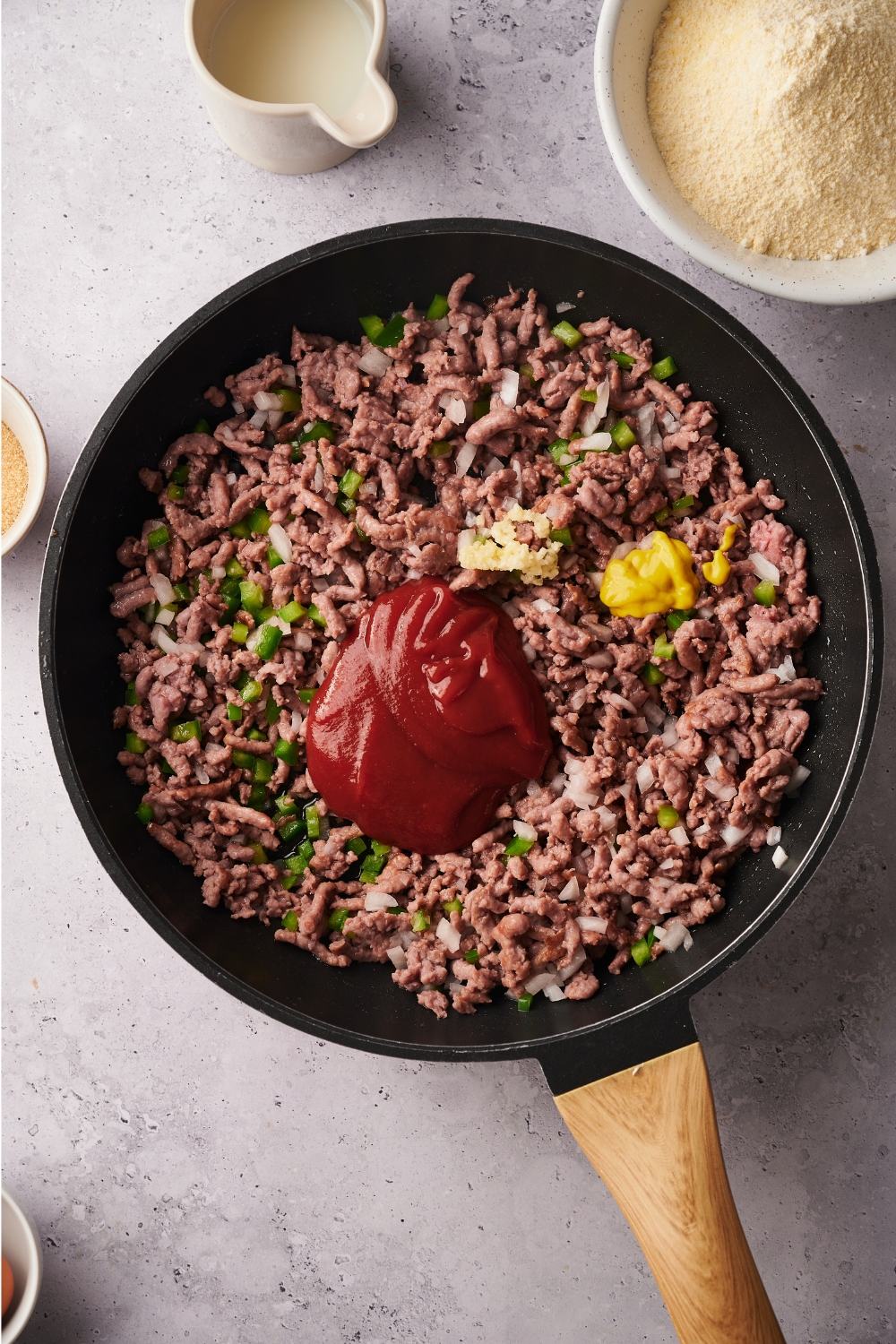 A skillet filled with cooked ground beef, green peppers, onion, minced garlic, yellow mustard, and ketchup.