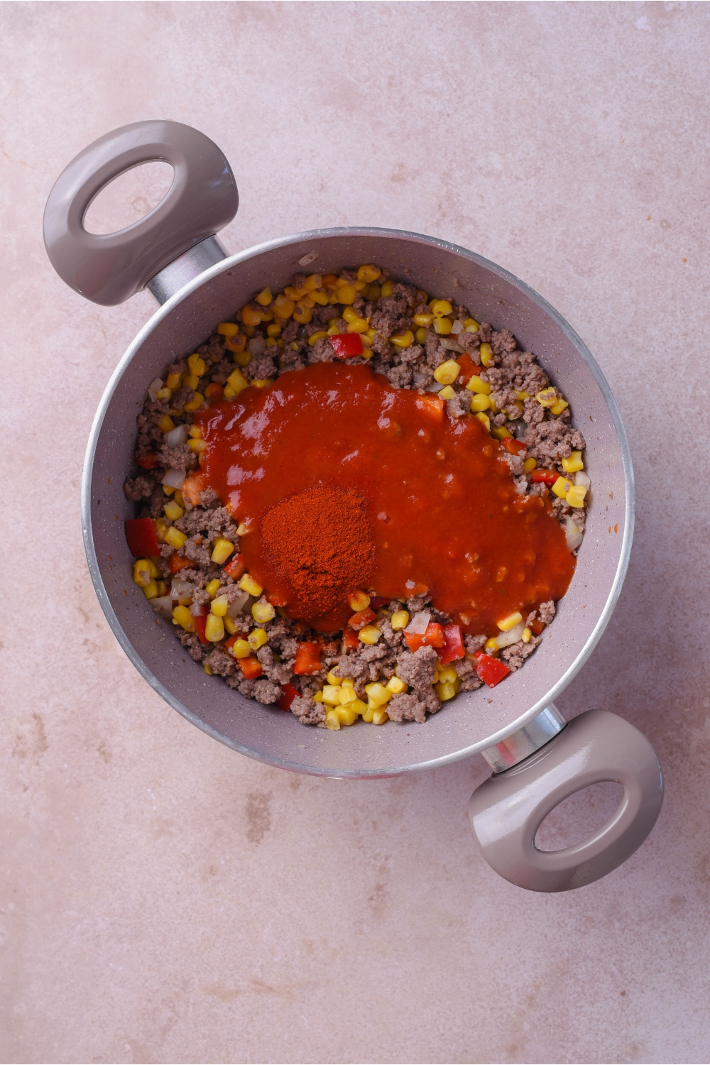 A pot filled with ground beef mixed with corn, red peppers, and onions, with red sauce and paprika poured on top but not yet mixed together.