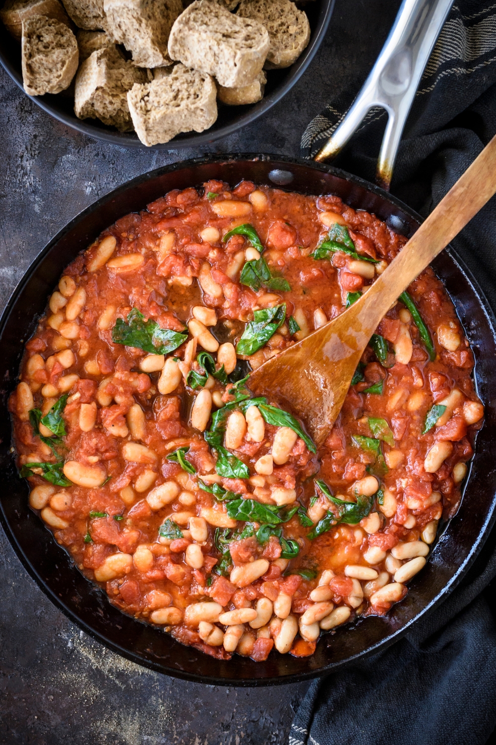 A skillet filled with tomato sauce, spinach, and white beans. There is a wooden spoon in the skillet.