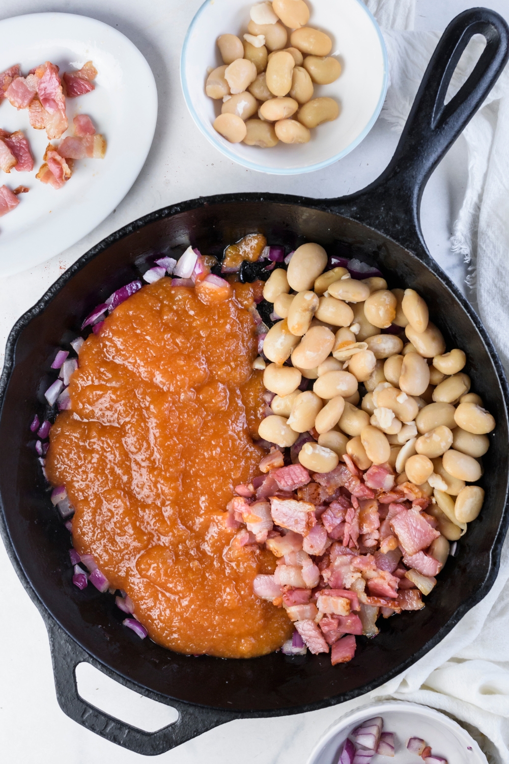A black skillet filled with butter beans, cooked bacon, diced onion, and a sauce mixture all added but not yet mixed together.