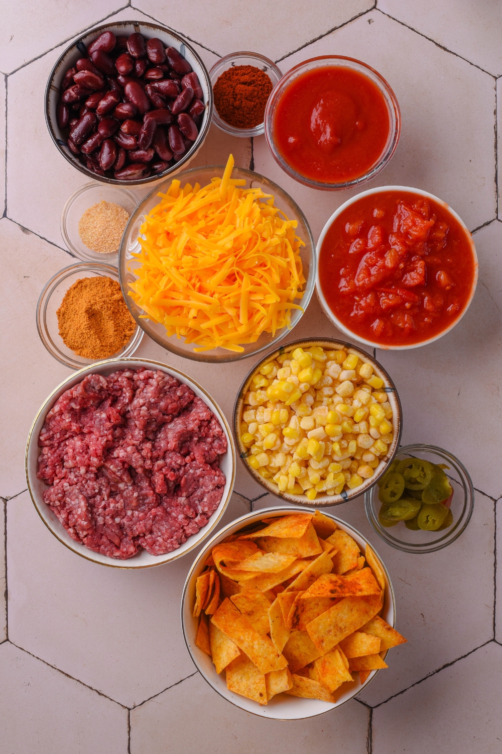 An assortment of ingredients including bowls of raw ground beef, shredded cheese, Fritos corn chips, corn, tomato sauce, beans, jalapeños, and spices.