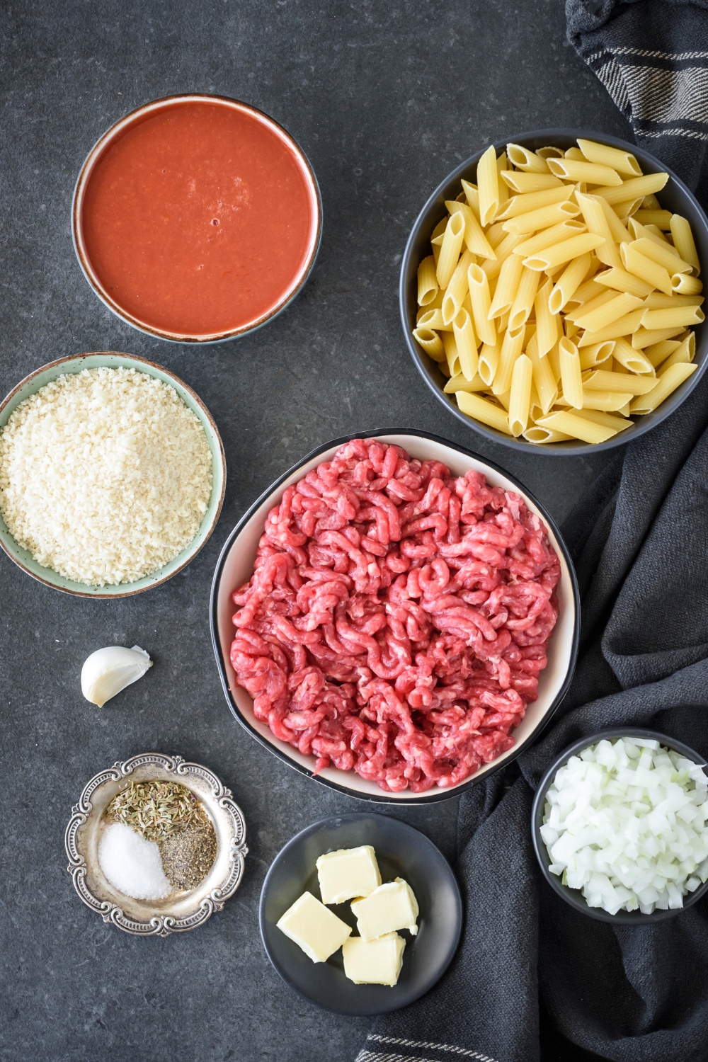 An assortment of ingredients including bowls of tomato sauce, dried pasta, raw ground beef, bread crumbs, diced onion, herbs, and a single garlic clove.