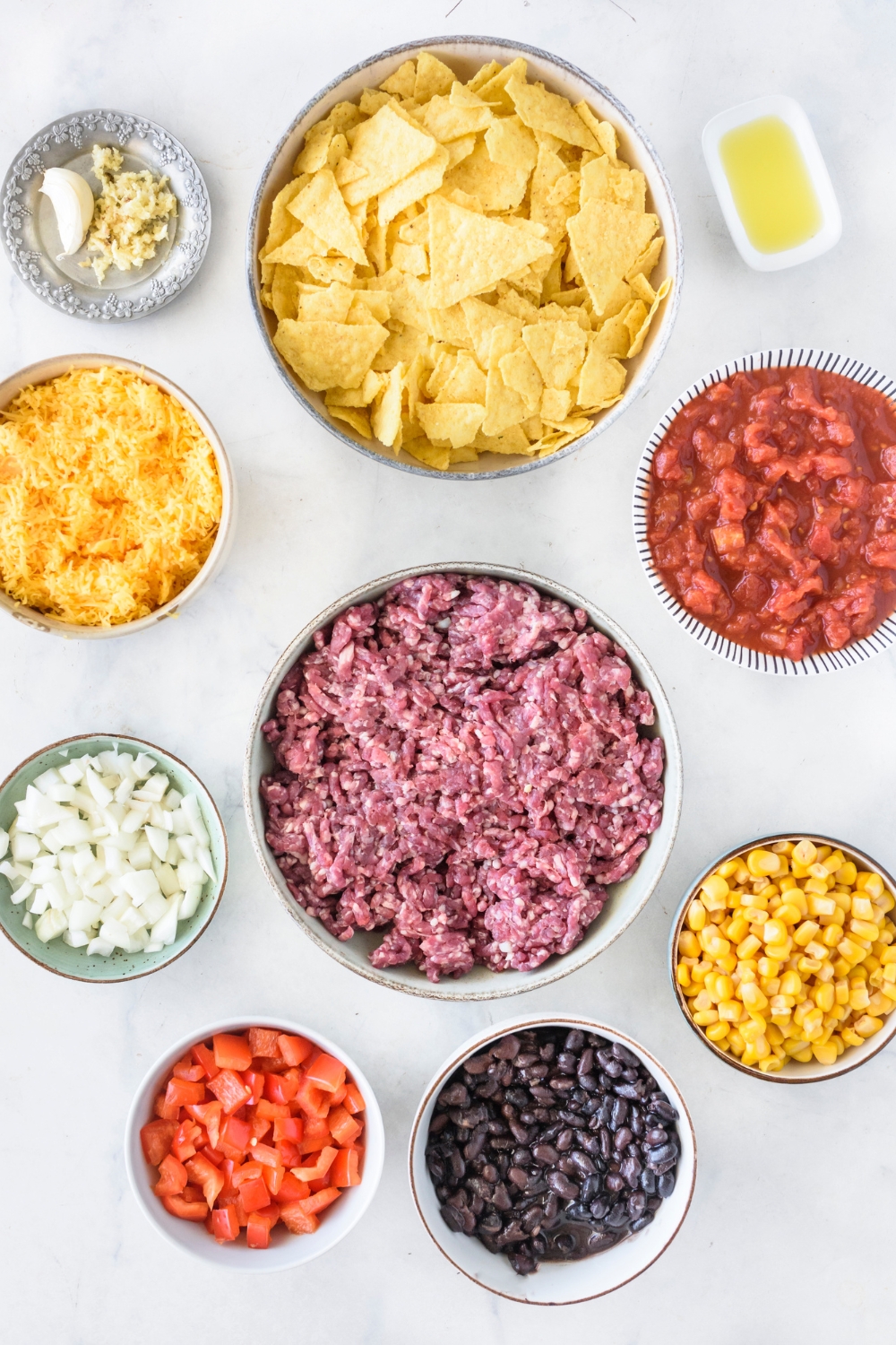 An assortment of ingredients including bowls of crushed tortilla chips, raw ground beef, diced red peppers, diced onion, shredded cheese, salsa, corn, and black beans.