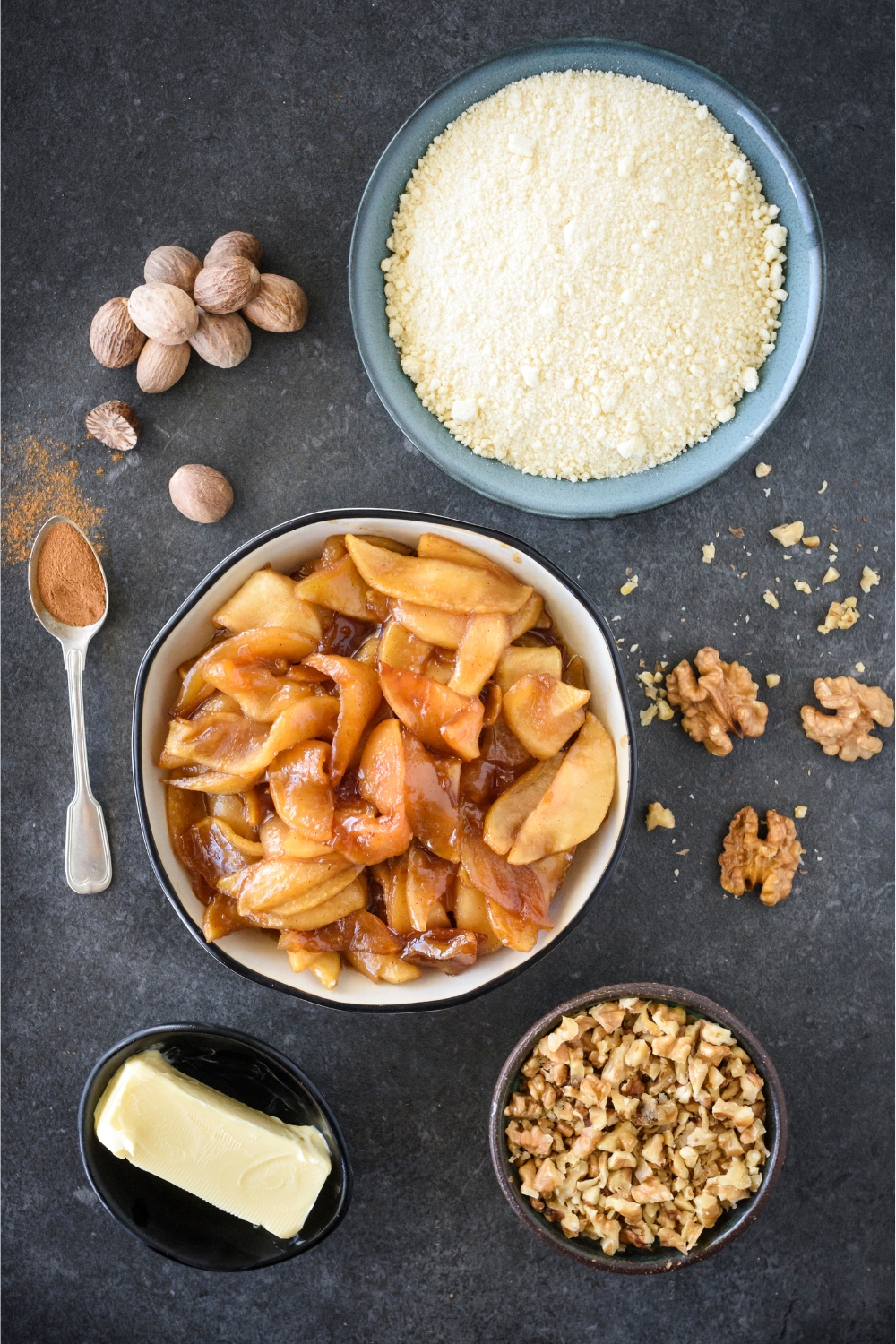 An assortment of ingredients including bowls of apple pie filling, cake mix, a butter stick, chopped nuts, a spoonful of cinnamon and a pile of nutmeg.