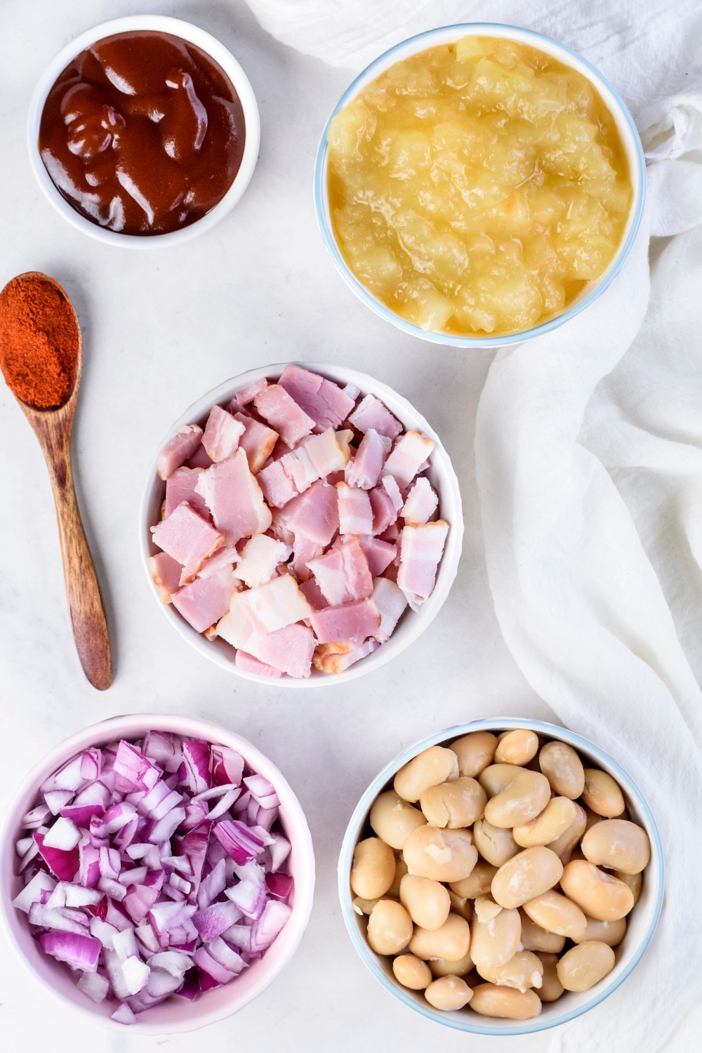 An assortment of ingredients including bowls of butter beans, diced onion, raw ham, barbecue sauce, apple pie filling, and a spoon of paprika.