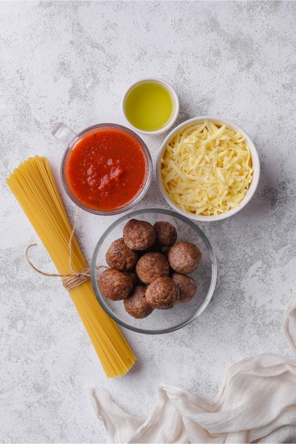 An assortment of ingredients including bowls of red sauce, oil, shredded cheese, meatballs, and a bundle of spaghetti noodles.