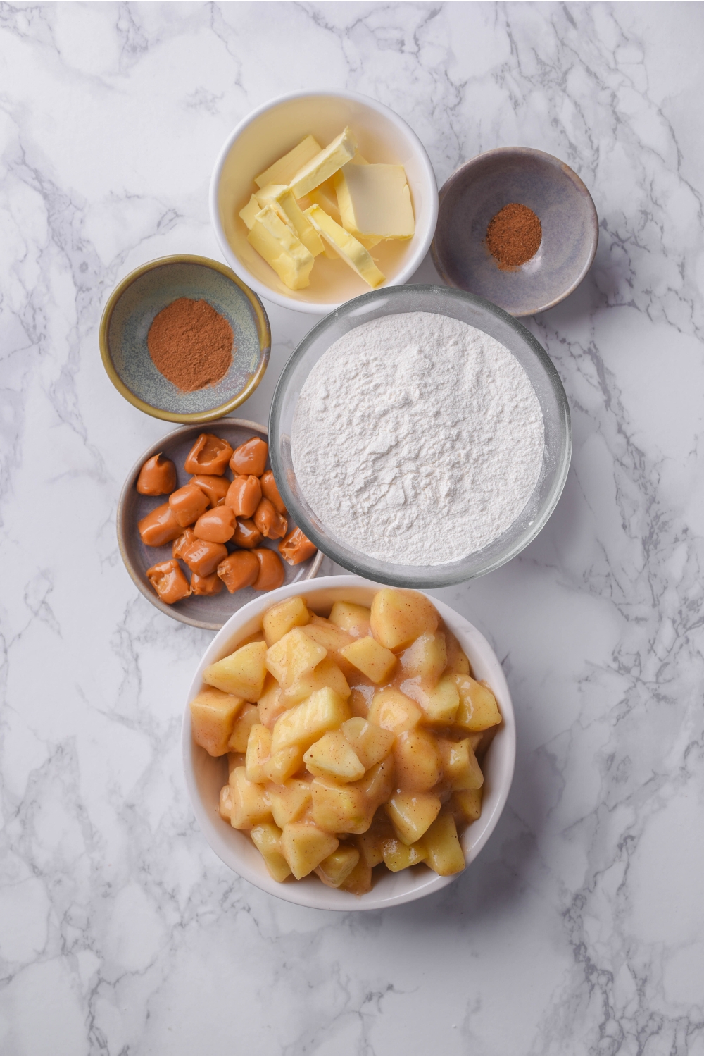 An assortment of ingredients including bowls of flour, apple pie filling, caramel candies, butter pats, cinnamon, and nutmeg.