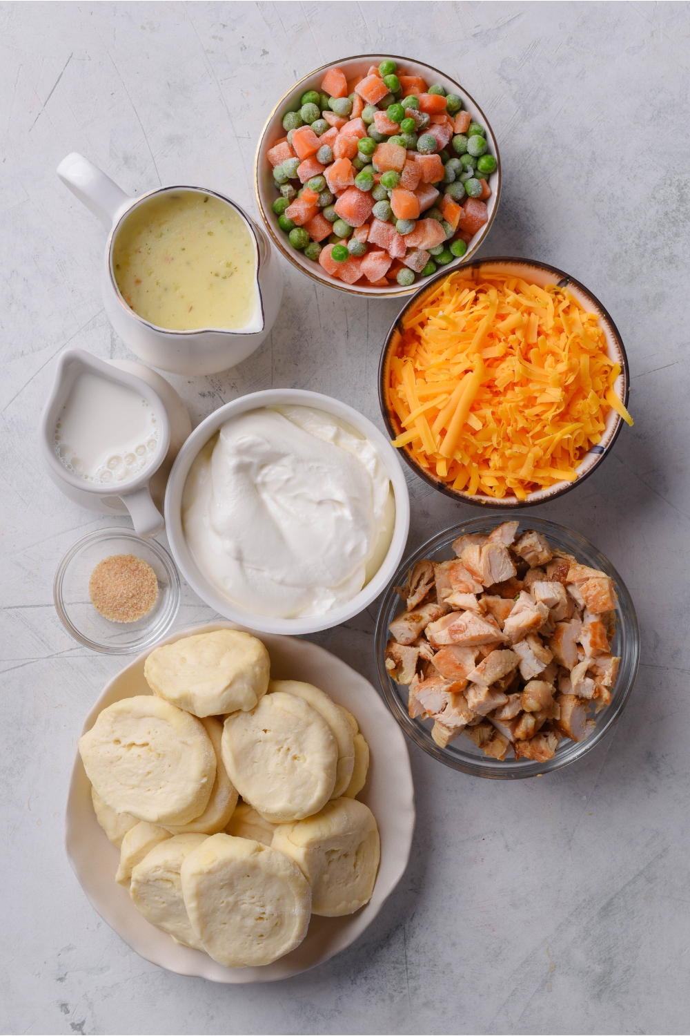 An assortment of ingredients including bowls of peas and carrots, shredded cheese, sour cream, diced chicken, biscuit dough slices, garlic powder, and pitchers of milk and cream of chicken soup.