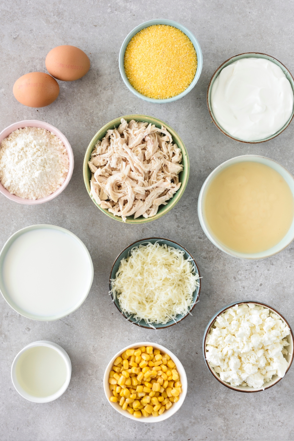 An assortment of ingredients including bowls of cornmeal, shredded chicken, flour, shredded cheese, whole corn kernels, feta cheese, yogurt, and two eggs.