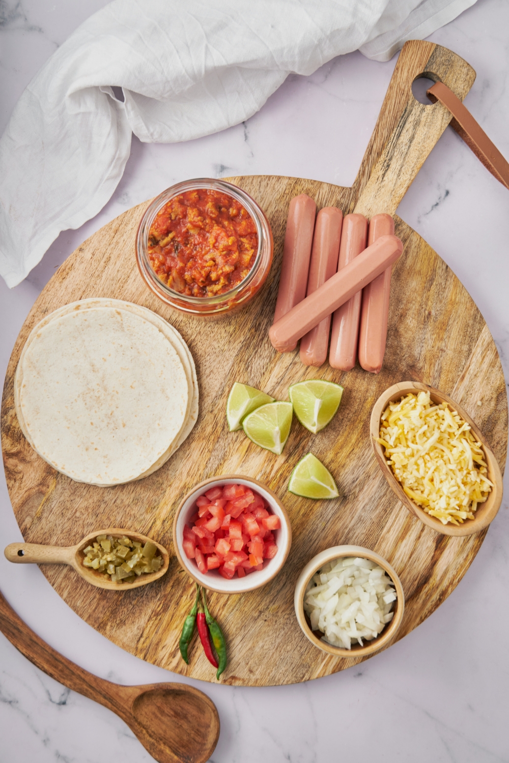 An assortment of ingredients including a stack of tortillas, a jar of chili, bowls of diced onion, diced tomato, shredded cheese, lime wedges, and hot dogs.