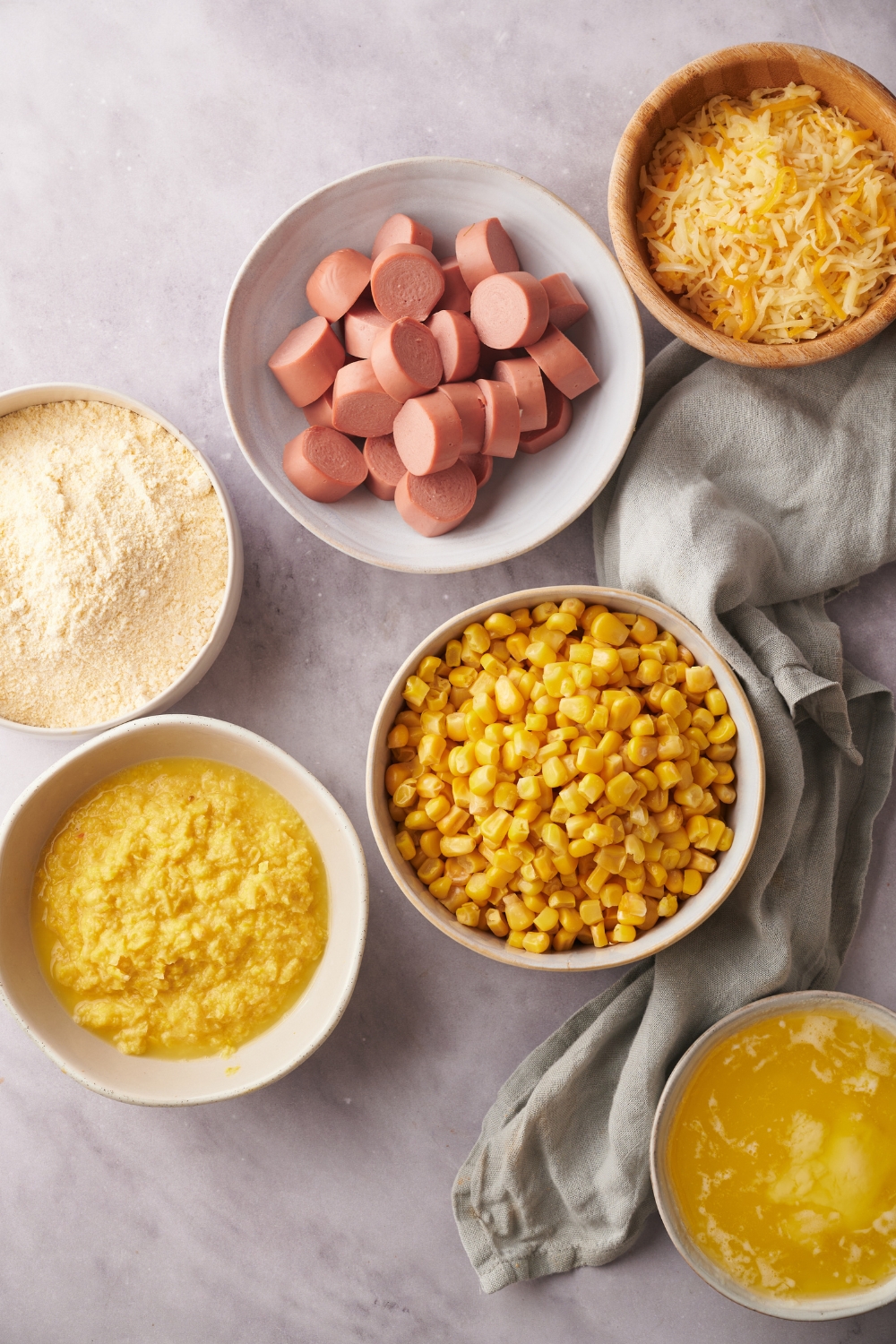 An assortment of ingredients including bowls of sliced hot dogs, corn, creamed corn, melted butter, shredded cheese, and a flour mixture.