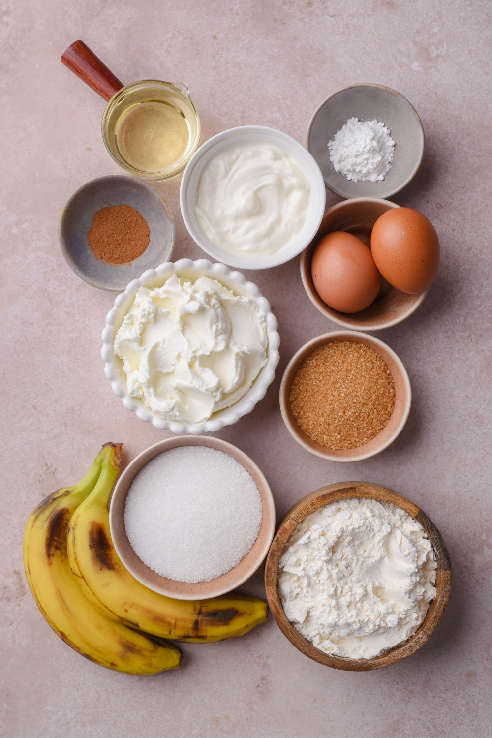 An assortment of ingredients including bowls of cream cheese, sugar, brown sugar, flour, baking soda, two eggs, oil, cinnamon, and two bananas.