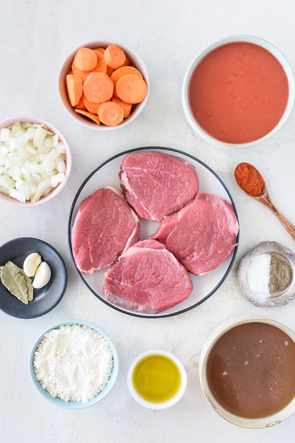 An assortment of ingredients including a plate of raw round steaks and bowls of carrots, oil. broth, tomato sauce, diced onion, salt and pepper, garlic and bay leaves, and a spoon of paprika.