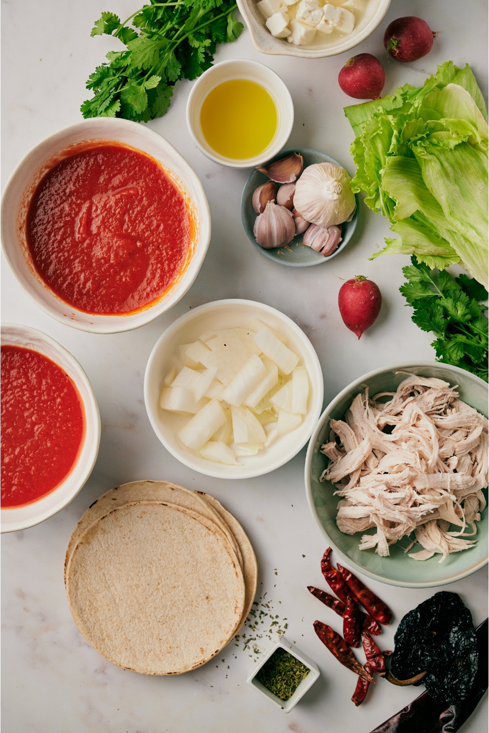 An assortment of ingredients including bowls of shredded chicken, tomato sauce, diced onion, oil, garlic cloves, a stack of tortillas, and a pile of dried peppers.