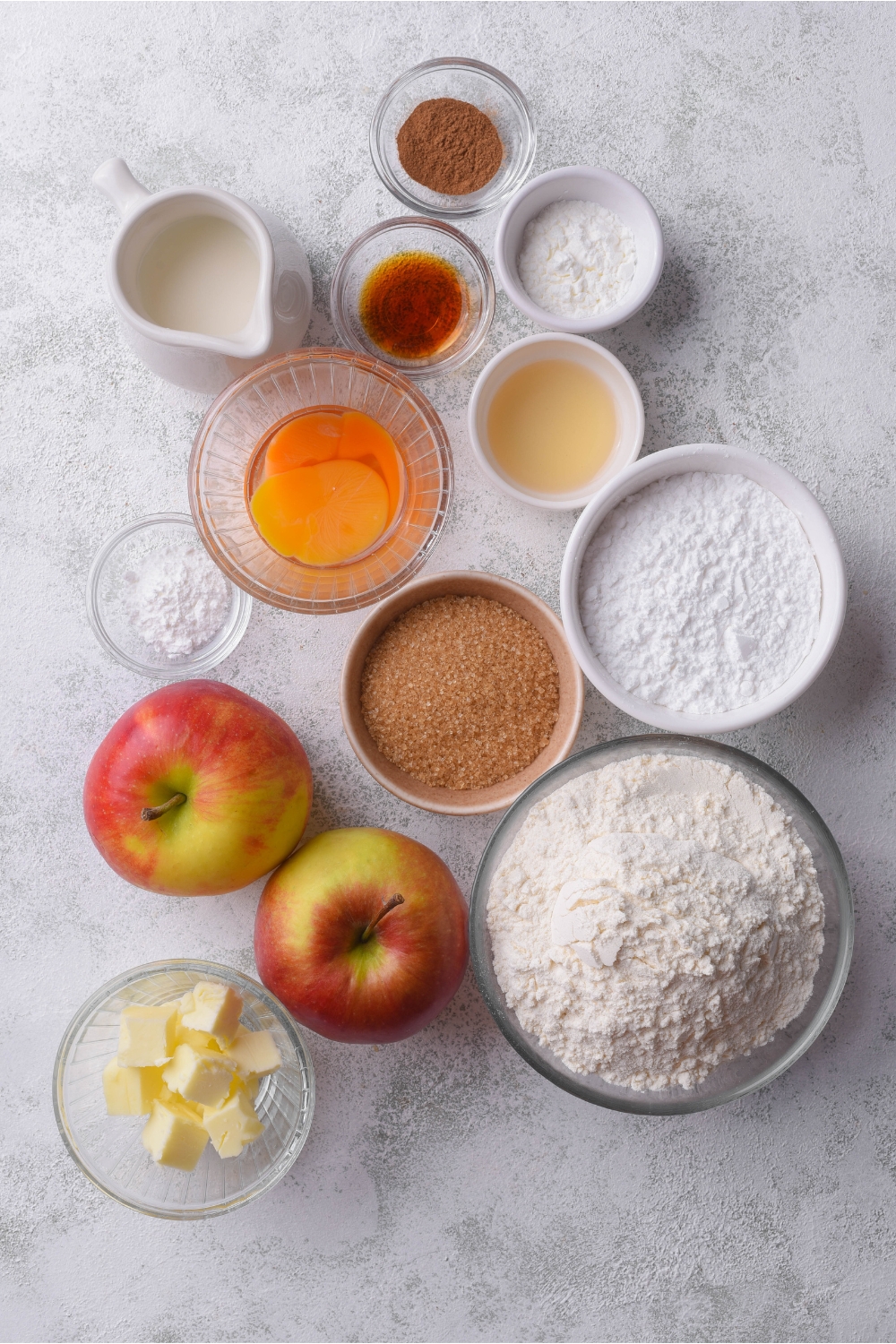 An assortment of ingredients including bowls of flour, powdered sugar, vanilla extract, brown sugar, butter cubes, a beaten egg, a jar of milk, and two apples.