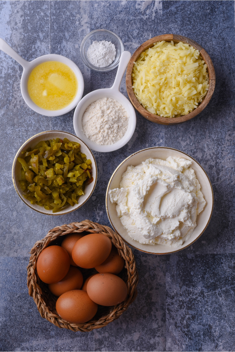 An assortment of ingredients including bowls of melted butter, diced green chilis, shredded cheese, flour, cottage cheese, and eggs.