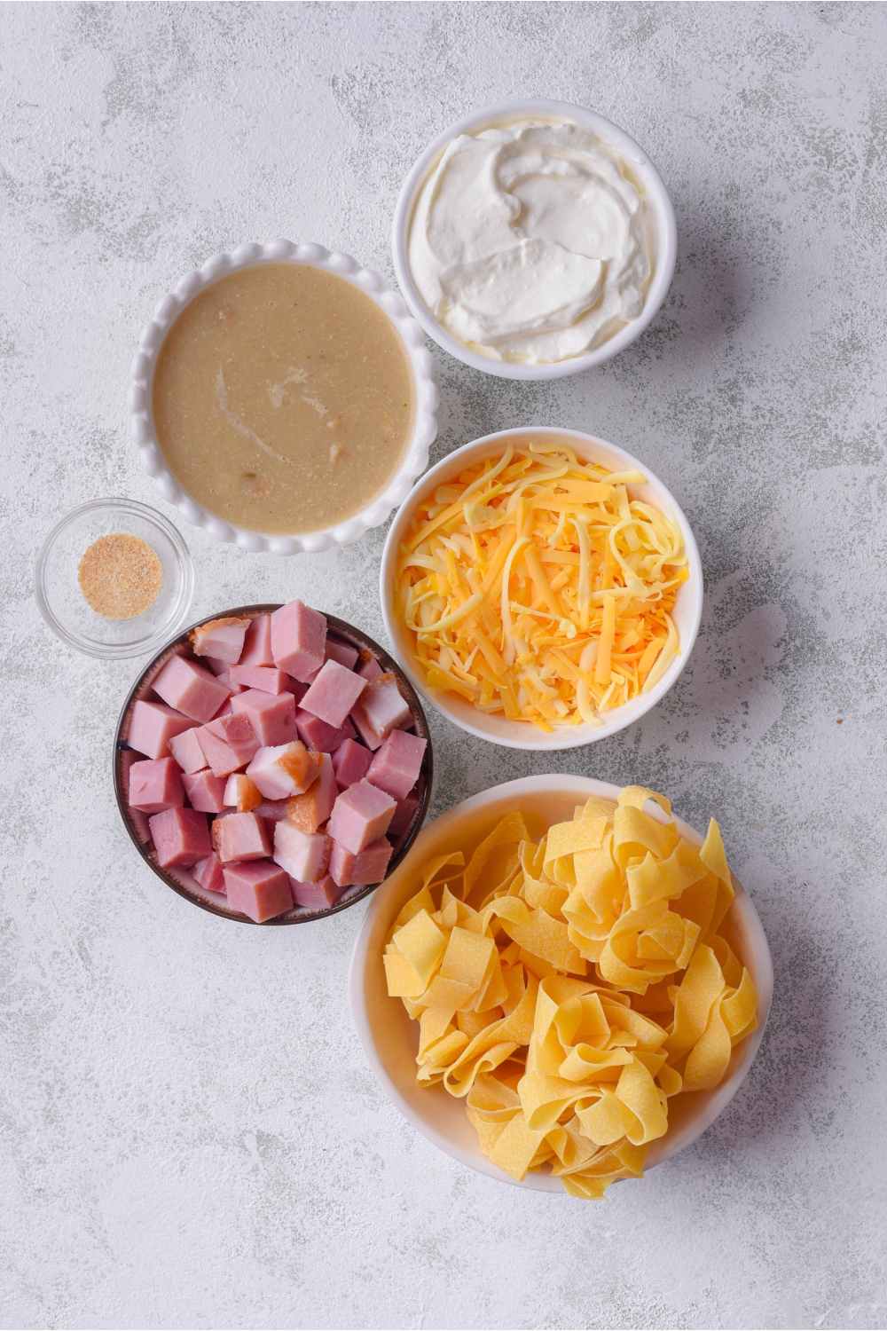 An assortment of ingredients including bowls of dried egg noodle nests, diced ham, shredded cheese, cream of mushroom soup, and sour cream.