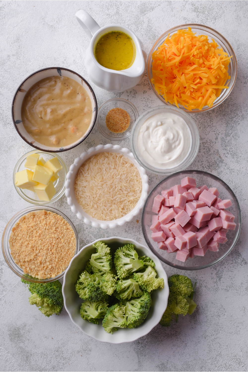 An assortment of ingredients including bowls of shredded cheese, broccoli, diced ham, crushed crackers, condensed soup, butter, and spices.