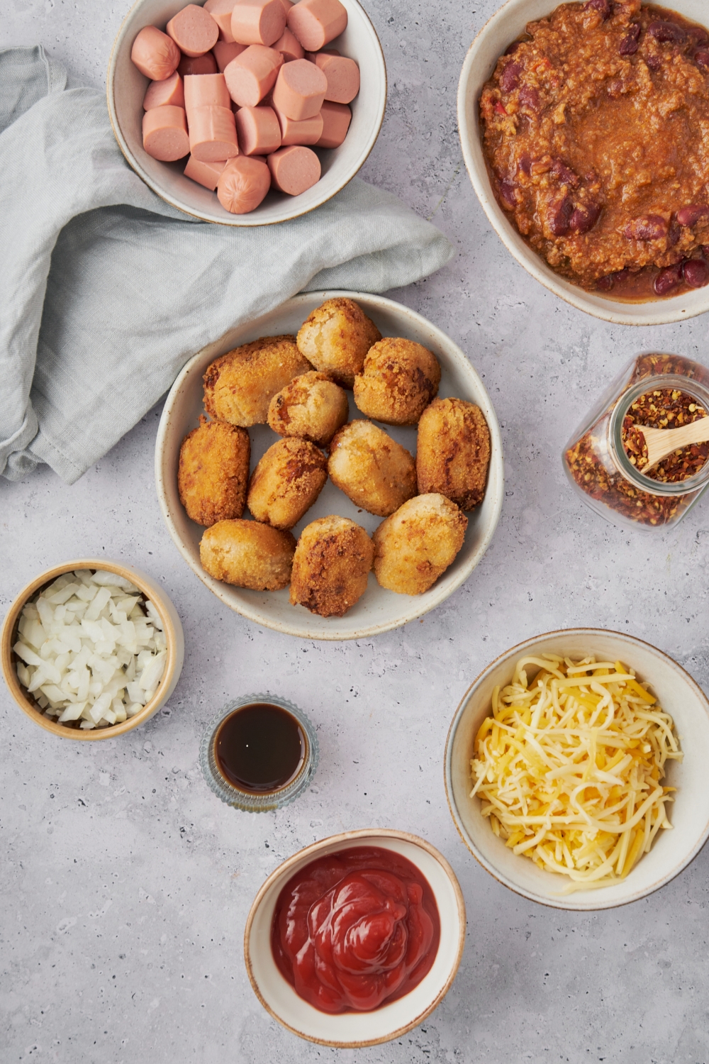 An assortment of ingredients including bowls of tater tots, chili, shredded cheese, ketchup, hot dog slices, diced onion, red pepper flakes, and Worcestershire sauce.