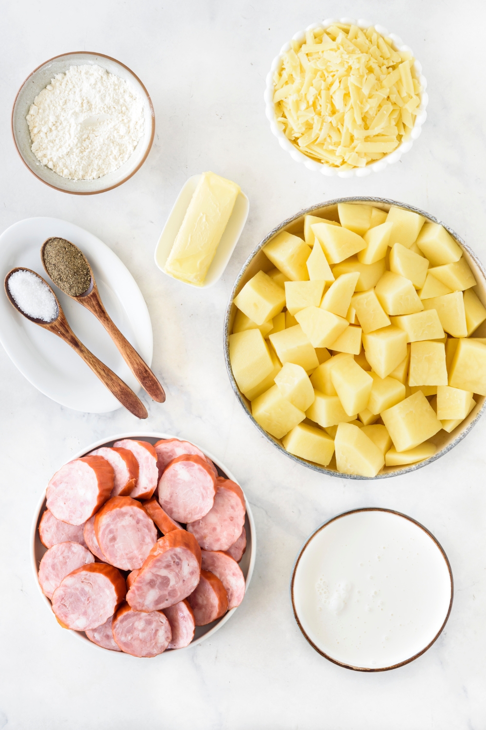 An assortment of ingredients including bowls of cubed potatoes, sliced sausages, shredded cheese, flour, butter, milk, and spoons of salt and pepper.