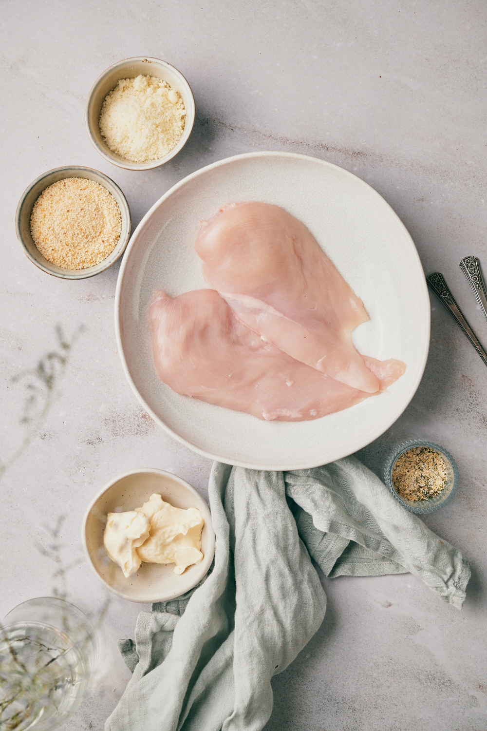 An assortment of ingredients including two raw chicken breasts on a plate next to bowls of breadcrumbs, parmesan cheese, mayonnaise, and seasonings.