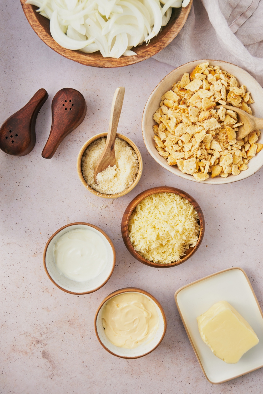 An assortment of ingredients including bowls of cracker crumbs, shredded cheese, mayonnaise, sour cream, butter, and sliced onions.