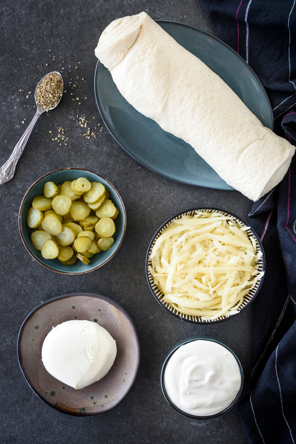 An assortment of ingredients including bowls of sliced pickles, shredded cheese, garlic sauce, a ball of fresh mozzarella, and a roll of pizza dough.