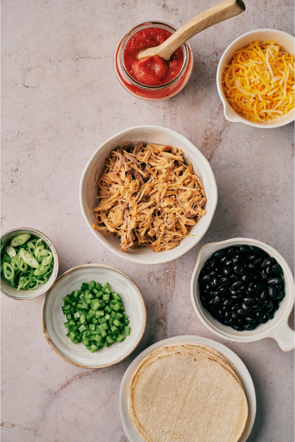 An assortment of ingredients including bowls of salsa, shredded cheese, pulled pork, black beans, diced bell pepper, green onion, and a plate of tortillas.