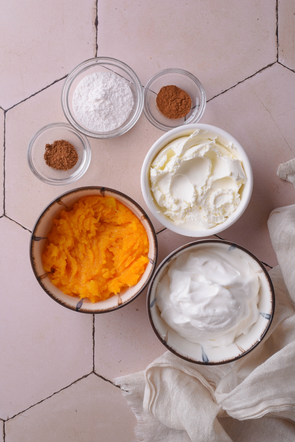 An assortment of ingredients including bowls of whipped cream, pumpkin puree, cream cheese, and spices.