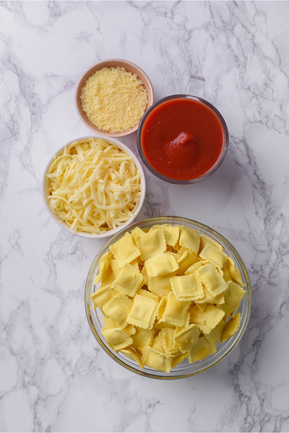 Bowls of parmesan cheese, marinara sauce, shredded mozzarella cheese, and mini raviolis.