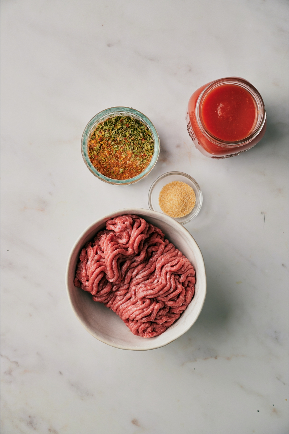 An assortment of ingredients including a bowl of raw ground beef, a jar of salsa, a bowl of taco seasoning, and a bowl of garlic powder.