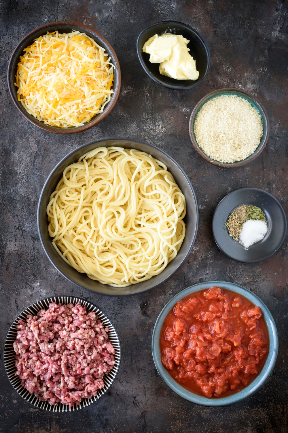 An assortment of ingredients including bowls of spaghetti, raw ground beef, tomato sauce, herbs, shredded cheese, butter, and bread crumbs.