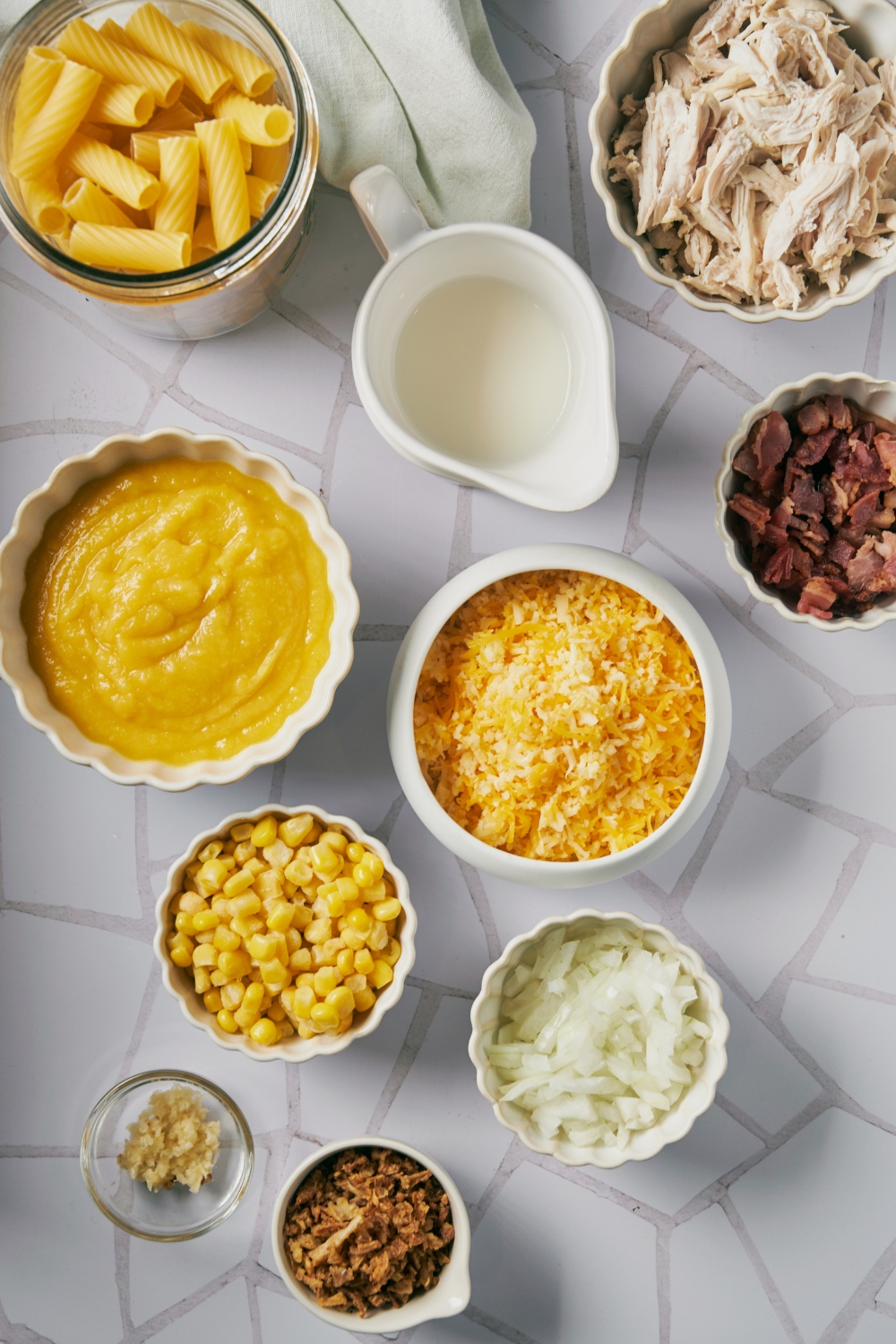 An assortment of ingredients including bowls of shredded cheese, diced onion, bacon, shredded chicken, dried pasta, fresh corn, condensed soup, garlic, and french fried onions.