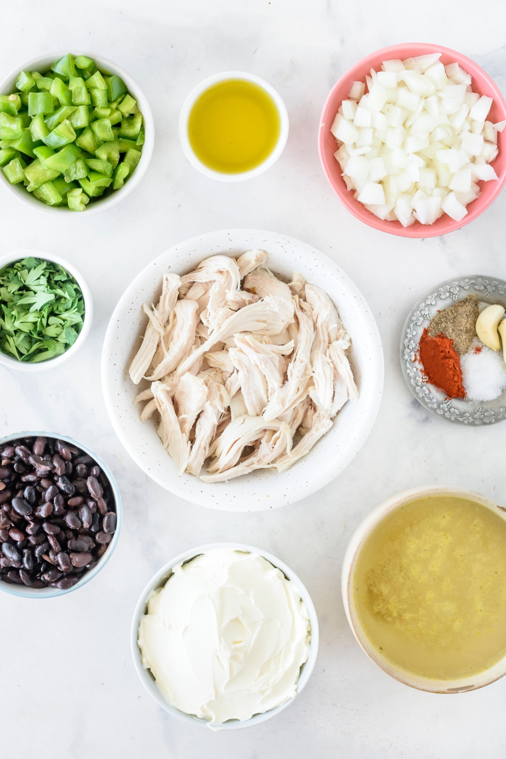 An assortment of ingredients including bowls of shredded chicken, diced onion, diced bell pepper, black beans, chicken broth, cream cheese, and spices.