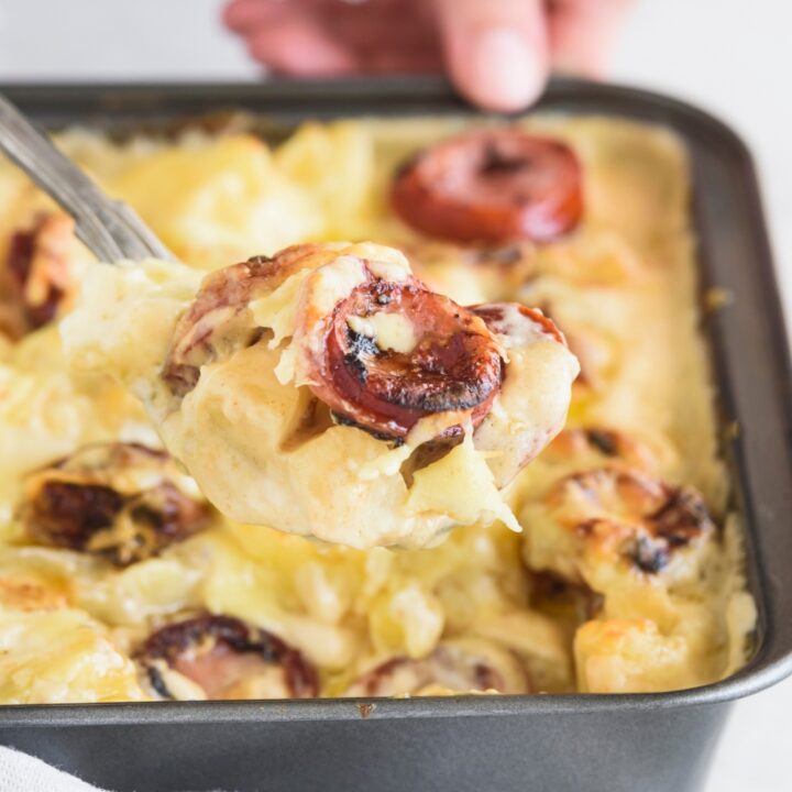 A serving spoon holding a scoop of sausage and potato casserole above a baking dish filled with more casserole.