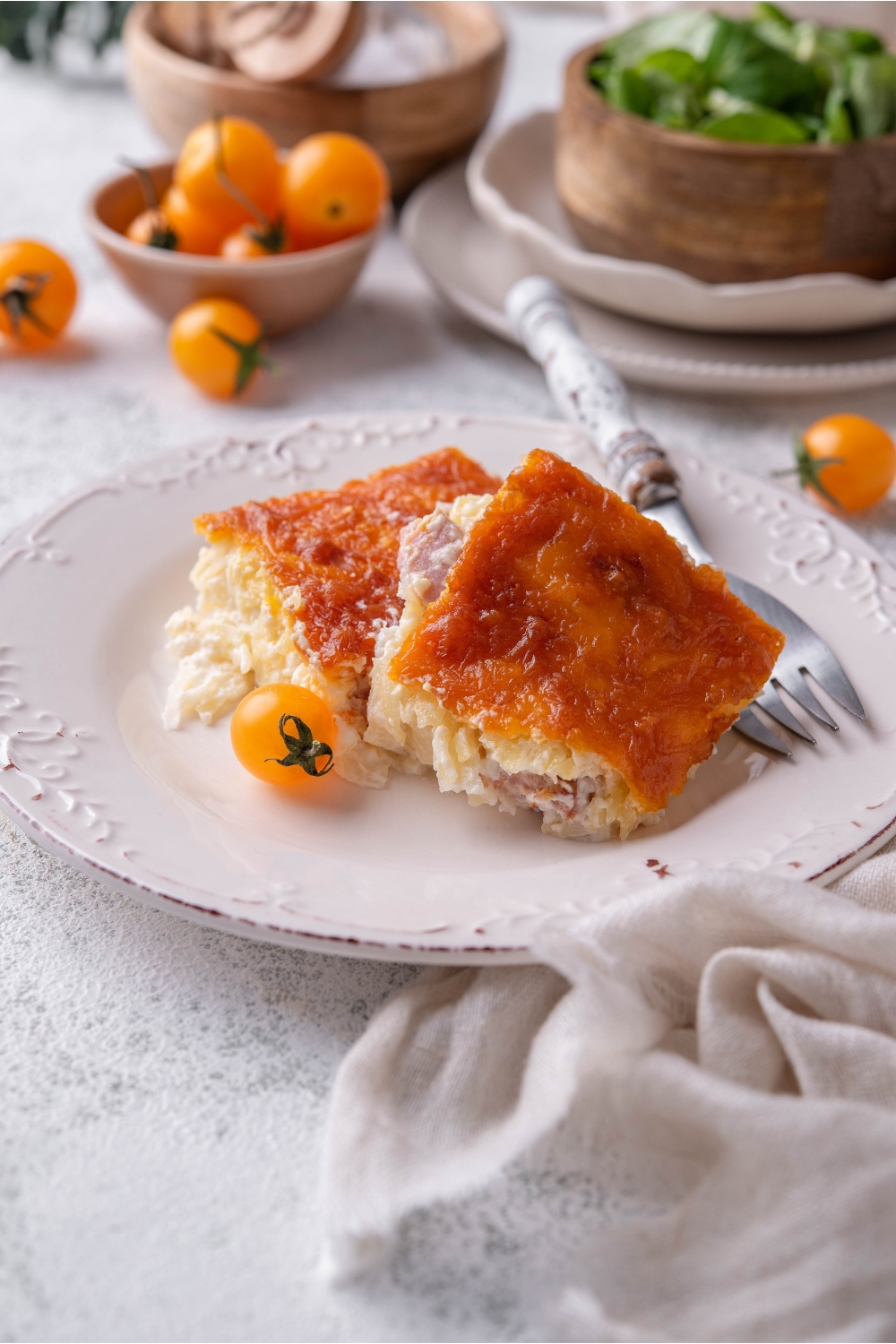 Two square servings of ham and hash brown casserole layered on top of each other with a cherry tomato and a fork on the plate next to the casserole.