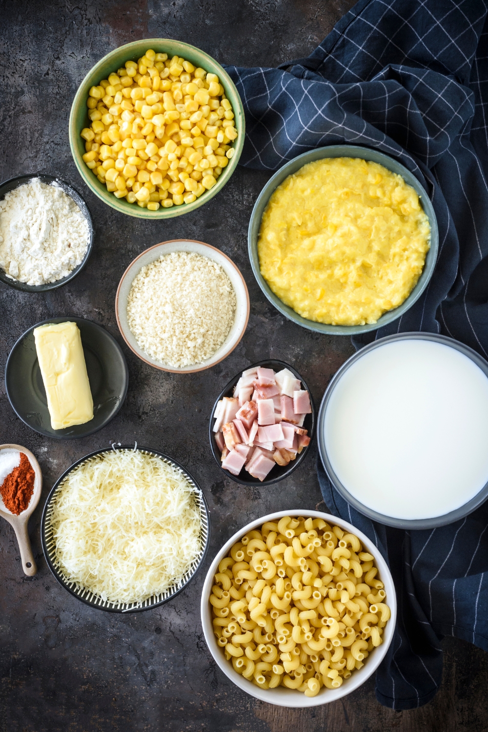 A countertop with multiple bowls with cooked pasta, corn, creamed corn, milk, cheese, butter, bacon, flour, and seasonings.