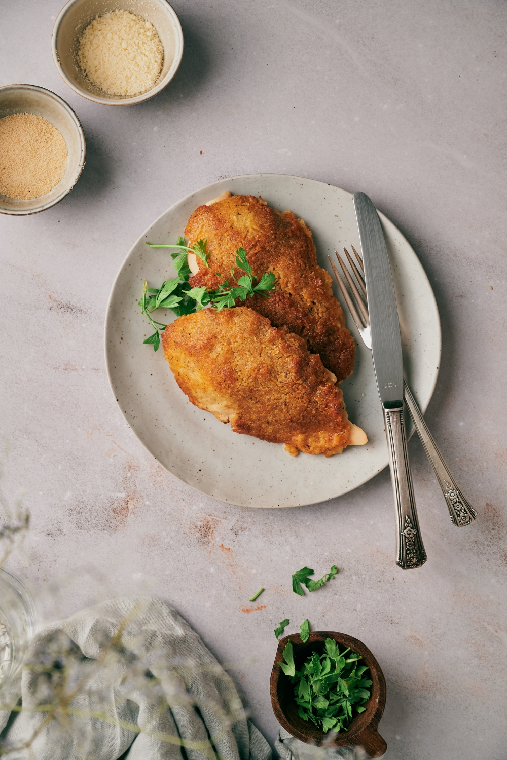 Two freshly baked and breaded chicken breasts on a plate, garnished with fresh herbs with a set of silverware on the plate next to the chicken.