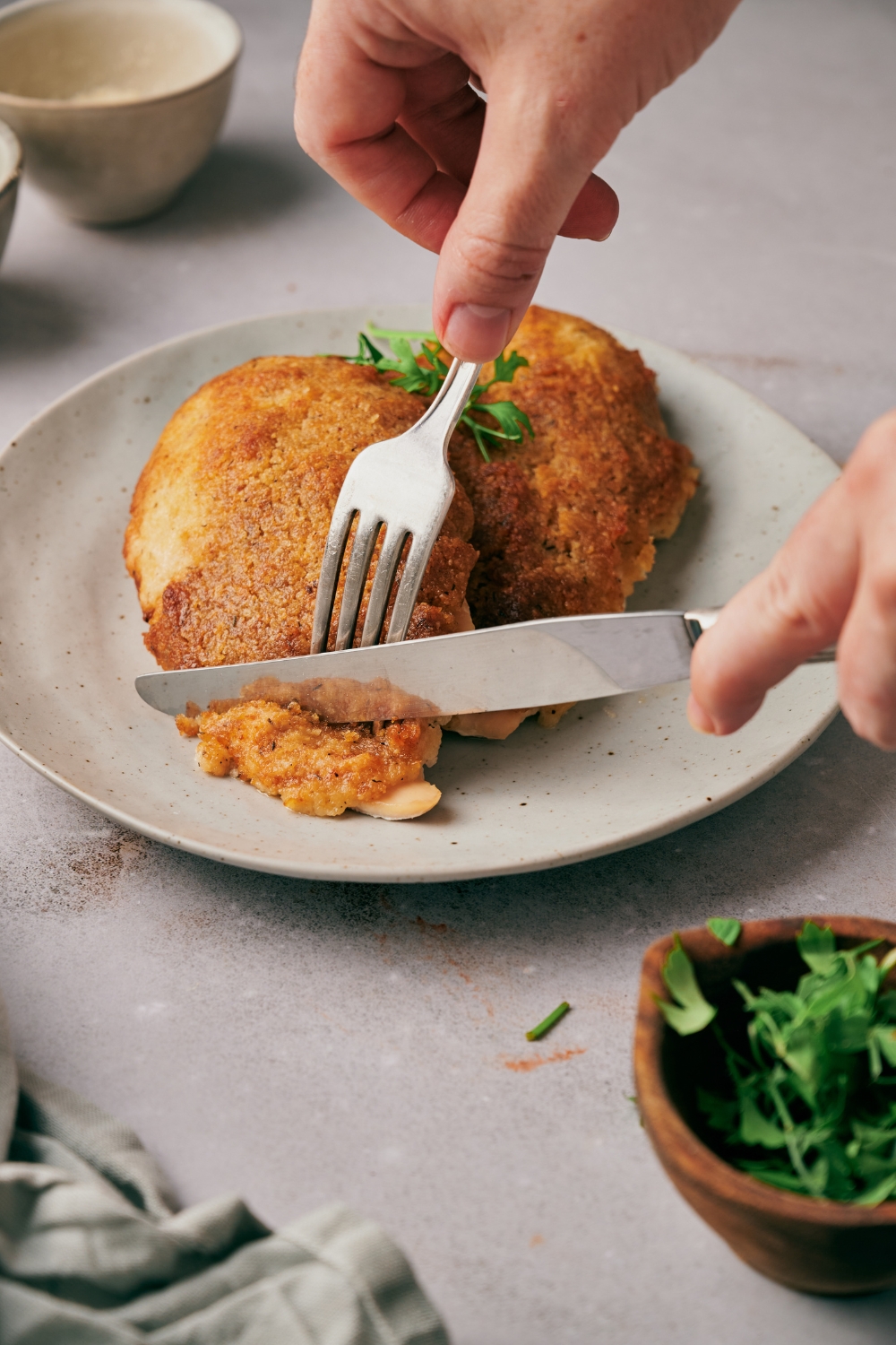 A hand using a fork and knife to slice a piece of baked chicken covered in crispy breading.