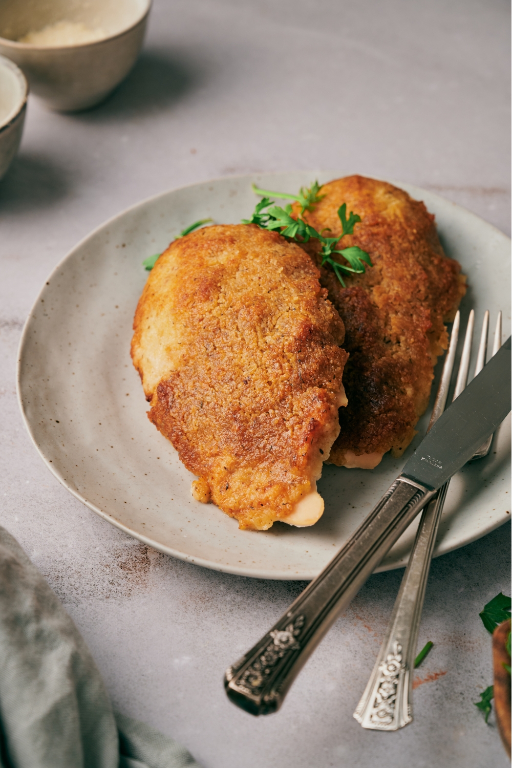 Two freshly baked and breaded chicken breasts on a plate, garnished with fresh herbs with a set of silverware on the plate next to the chicken.