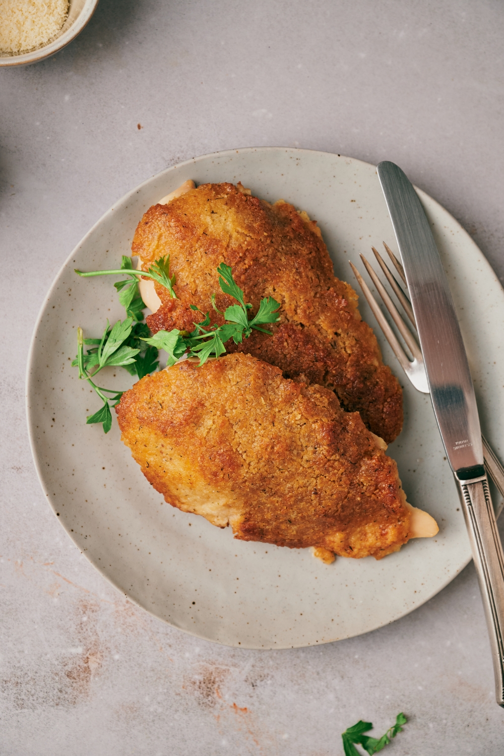 Two freshly baked and breaded chicken breasts on a plate, garnished with fresh herbs with a set of silverware on the plate next to the chicken.