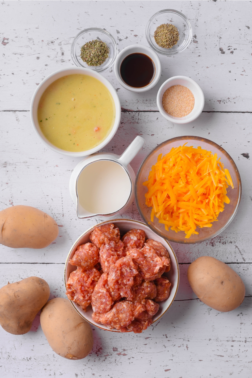 An assortment of ingredients including bowls of raw sausage, shredded cheese, cream of mushroom soup, spices, herbs, a pitcher of milk, and four potatoes on the counter.