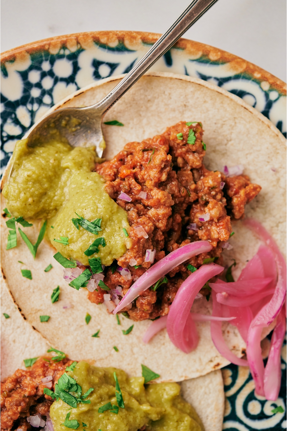 A taco topped with seasoned taco meat, diced red onion, pickled red onion, fresh herbs, and a spoonful of green salsa with a spoon still on the plate.