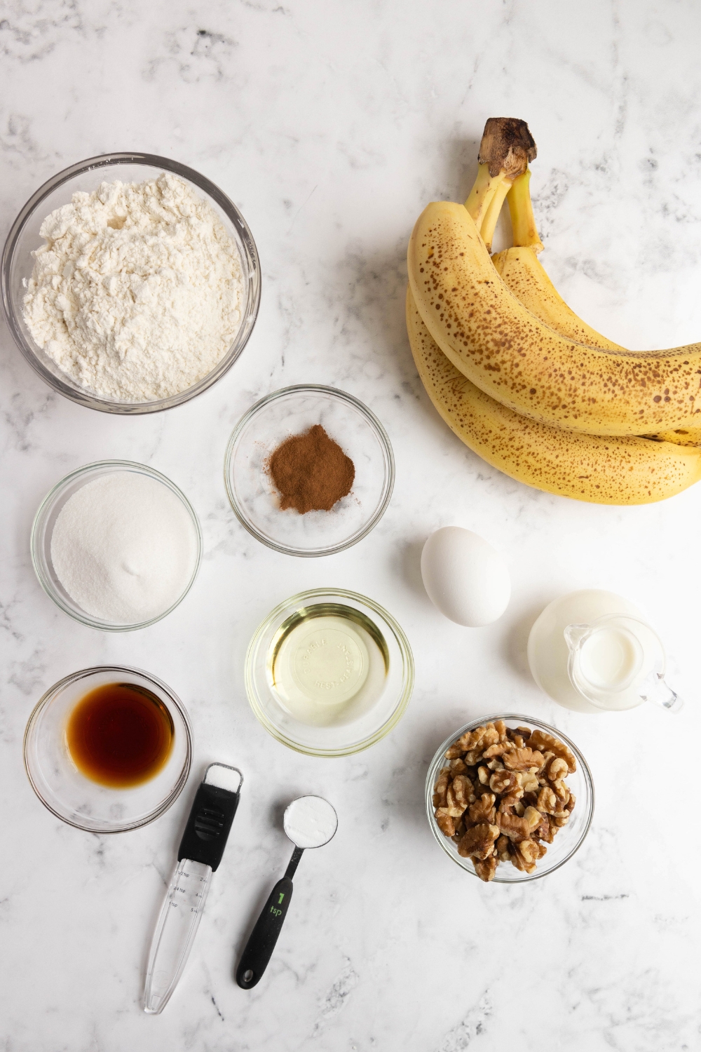 A countertop with flour, sugar, cinnamon, vanilla, oil, buttermilk, chopped walnuts, salt, baking soda, an egg, and bananas in separate containers and bowl.