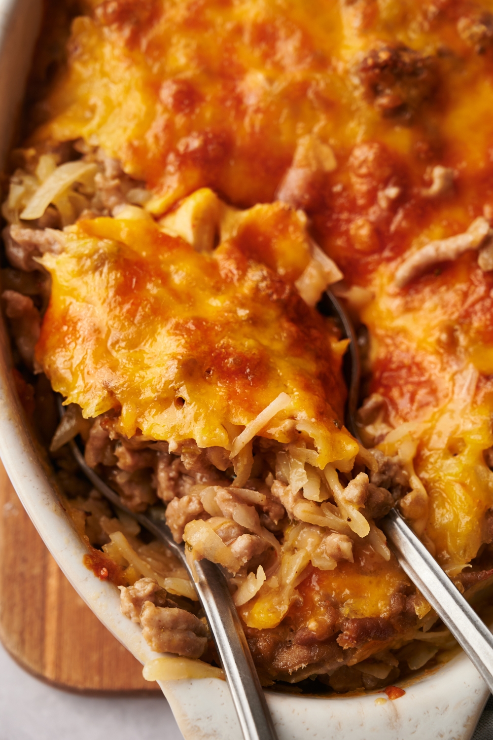 Close up of hash brown and ground beef casserole in a baking dish covered with melted cheese and two spoons are in the casserole.