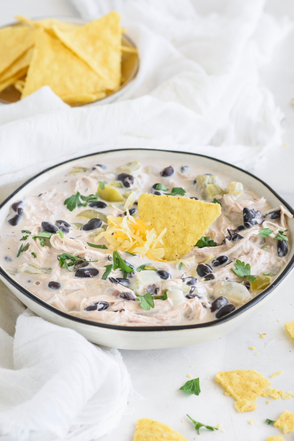 A bowl of white chicken chili with parsley and black beans in the chili and shredded cheese and a corn chip are garnished on top.